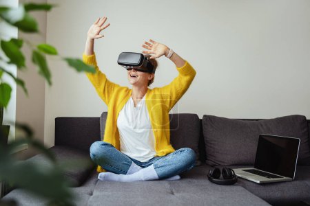 Téléchargez les photos : Jeune femme excitée assise sur le canapé et utilisant un casque de réalité virtuelle à la maison - en image libre de droit
