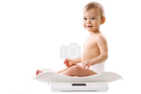 Foto de Medida de peso de un niño adorable en pañal sentado en escamas sobre fondo blanco. - Imagen libre de derechos