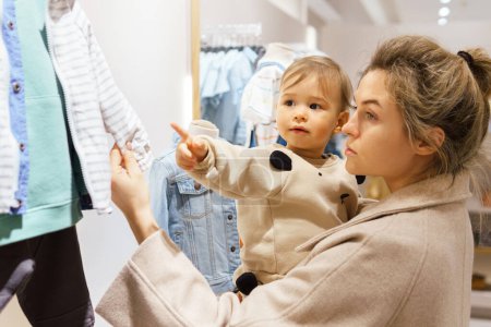 Foto de Mujer joven con su hijo bebé eligiendo ropa en la tienda de ropa - Imagen libre de derechos