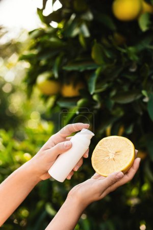 Téléchargez les photos : Femme tient une bouteille blanche de produit de soins de la peau et la moitié d'un citron juteux dans ses mains sur fond de citronnier flou. - en image libre de droit