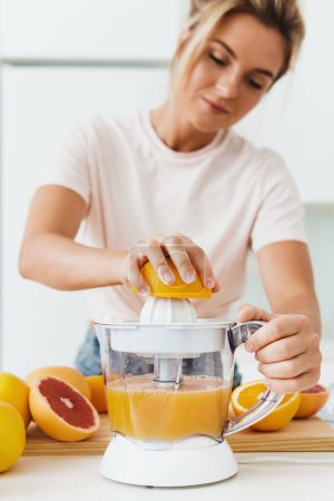 Photo for Young beautiful woman preparing orange juice with modern citrus juicer machine at home - Royalty Free Image