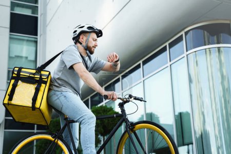 Foto de Joven entrega de comida express mensajero montar en bicicleta con bolsa aislante a sus espaldas está mirando su reloj. - Imagen libre de derechos
