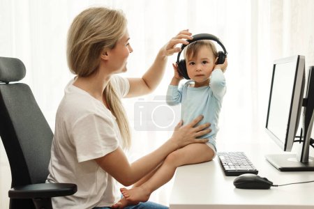 Foto de Joven madre freelancer pone sus auriculares inalámbricos en su pequeño hijo sentado en el escritorio de su oficina en casa. - Imagen libre de derechos