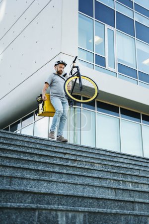 Foto de Joven mensajero de reparto de comida express con bolsa aislada detrás de su espalda está llevando su bicicleta por las escaleras. - Imagen libre de derechos