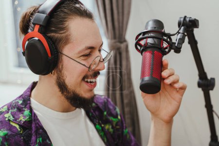 Téléchargez les photos : Jeune blogueur homme élégant utilisant un casque et un microphone à condensateur pendant le flux de podcast en ligne - en image libre de droit