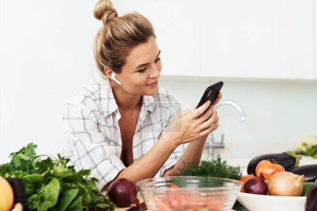Photo for Young pretty woman with wireless earbuds is using smartphone during cooking in modern white kitchen - Royalty Free Image