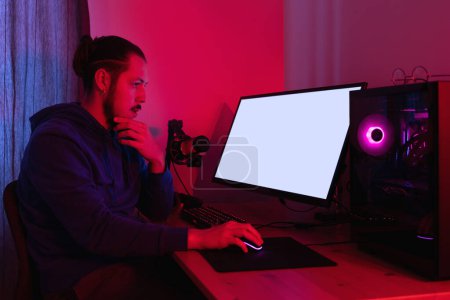 Foto de Hombre joven que trabaja en la PC moderna por la noche en la habitación con luz de neón. Pantalla de monitor en blanco para su diseño. - Imagen libre de derechos