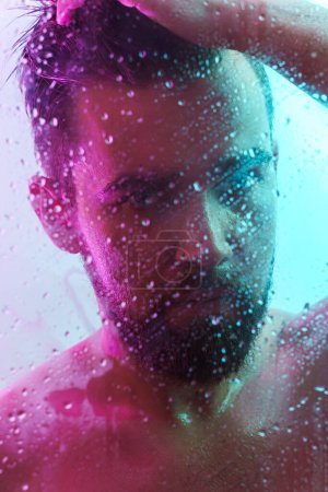 Téléchargez les photos : Portrait de beau jeune homme capturé à travers le verre mouillé dans la lumière au néon coloré - en image libre de droit