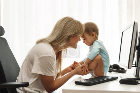 Téléchargez les photos : Jeune mère indépendante souriante joue avec son petit fils assis sur le bureau à son lieu de travail à la maison. - en image libre de droit