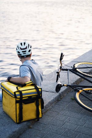 Téléchargez les photos : Jeune messager de livraison express est assis sur un quai et regardant l'eau calme de la rivière avec sac isolé et vélo à proximité. - en image libre de droit