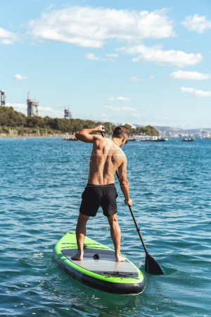 Photo for Young male surfer is riding a standup paddleboard and rowing with a paddle in ocean near the shore. - Royalty Free Image