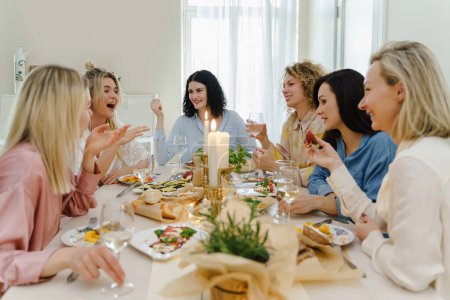 Foto de Jóvenes felices amigas bebiendo vino y riéndose en la mesa festiva servida con comida fresca y decorada con velas. - Imagen libre de derechos