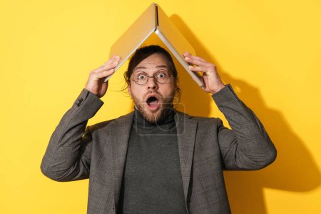 Foto de Shocked barbudo hombre sosteniendo ordenador portátil por encima de su cabeza en el fondo amarillo - Imagen libre de derechos