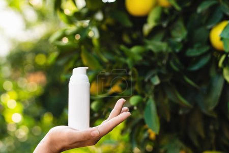 Téléchargez les photos : Femme tient une bouteille blanche de produit de soins de la peau dans sa main sur fond de citronnier flou. - en image libre de droit