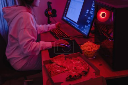 Photo for Young woman gamer or software developer sitting at the modern personal computer and eating junk food at night in room with neon lights - Royalty Free Image