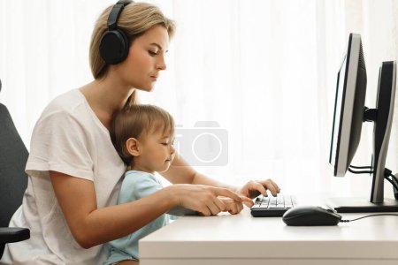 Foto de Joven madre freelancer está trabajando en la computadora en el lugar de trabajo de su oficina en casa con su pequeño hijo sentado en su regazo. - Imagen libre de derechos