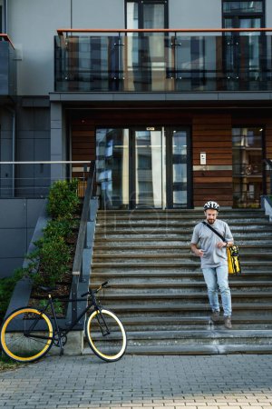 Photo for Young express food delivery courier with insulated bag behind his back is going down the stairs to his parked bicycle. - Royalty Free Image