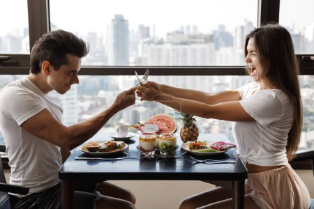 Téléchargez les photos : Jeune couple joyeux ludique se bat avec des couverts pendant le petit déjeuner - en image libre de droit