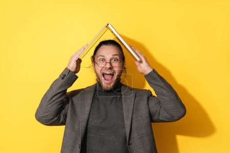 Foto de Joven hombre barbudo alegre sosteniendo ordenador portátil por encima de su cabeza sobre fondo amarillo - Imagen libre de derechos