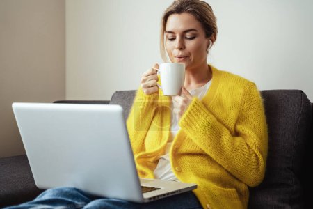 Photo for Young woman with cup of hot coffee sitting on the sofa and using laptop computer at home - Royalty Free Image