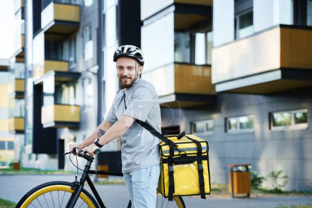 Photo for Young smiling express food delivery courier with an insulated bag behind his back is standing with a bicycle. - Royalty Free Image