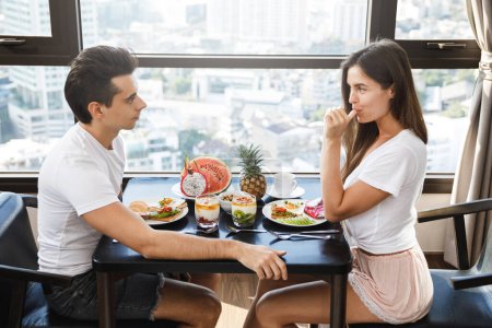 Téléchargez les photos : Jeune couple heureux petit déjeuner sain dans un appartement moderne avec de grandes fenêtres et vue sur la ville - en image libre de droit