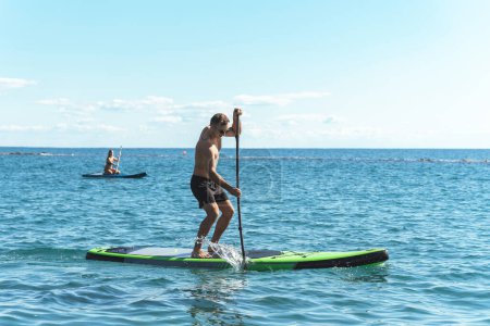 Photo for Young male surfer is riding a standup paddleboard and rowing with a paddle in an ocean. - Royalty Free Image