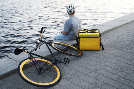 Photo for Young express delivery courier is sitting on a quay and looking at the quiet river water with insulated bag and bicycle nearby. - Royalty Free Image