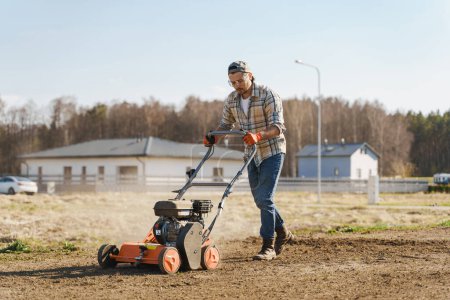 Téléchargez les photos : Jeune homme utilisant une machine d'aération pour scarifier et aérer la pelouse ou le pré - en image libre de droit