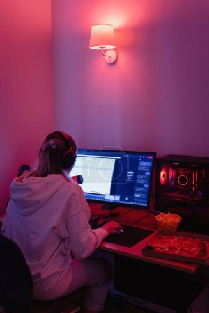 Young woman gamer or software developer sitting at the modern personal computer and eating junk food at night in room with neon lights Poster 653700416