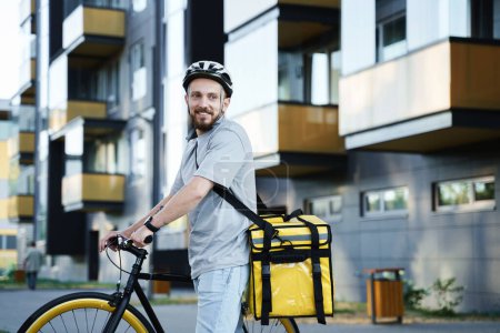 Foto de Joven sonriente mensajero de reparto de comida express con una bolsa aislada detrás de su espalda está de pie con una bicicleta. - Imagen libre de derechos
