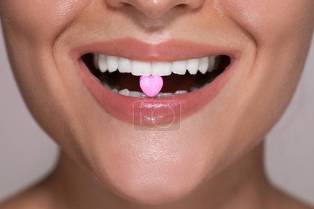 Foto de Primer plano de una parte inferior de una cara femenina joven con una píldora rosa en forma de corazón entre sus dientes. - Imagen libre de derechos