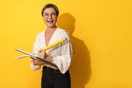 Téléchargez les photos : Joyeux jeune femme portant des lunettes tenant un gros crayon et un cahier sur fond jaune - en image libre de droit