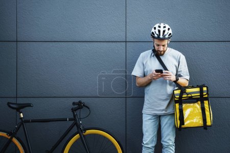Téléchargez les photos : Jeune messager express de livraison de nourriture avec sac isolé sur le côté regarde son téléphone près du vélo garé. - en image libre de droit