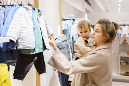 Téléchargez les photos : Jeune femme avec son bébé fils choisir des vêtements dans le magasin de vêtements - en image libre de droit