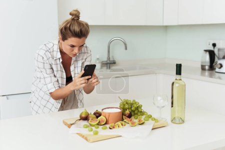 Téléchargez les photos : Jeune jolie femme prenant en photo son délicieux yaourt grec aux figues par son smartphone en cuisine blanche - en image libre de droit