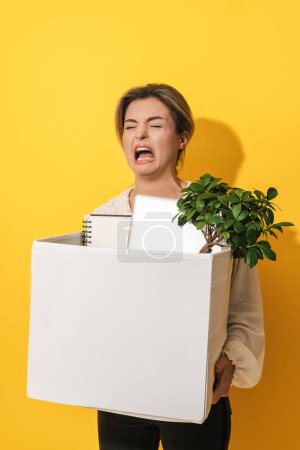 Foto de Joven mujer molesta caja de espera con artículos personales después de la renuncia al trabajo contra el fondo amarillo - Imagen libre de derechos