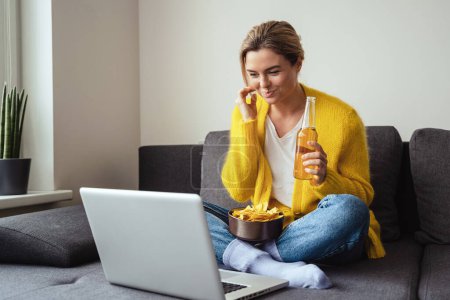 Téléchargez les photos : Jeune femme est assise sur le canapé à boire de la bière et manger des nachos tout en regardant un film sur son ordinateur portable à la maison - en image libre de droit