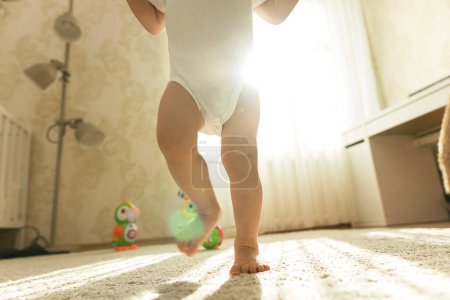 Téléchargez les photos : Petit garçon en body fait ses premiers pas sur un tapis recouvert de sol dans sa chambre. - en image libre de droit