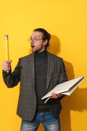 Foto de Hombre barbudo divertido con gafas sosteniendo lápiz grande y cuaderno sobre fondo amarillo - Imagen libre de derechos