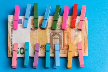 Gros plan de pinces à linge en bois colorées attachées à un billet de cinquante euros sur fond bleu.