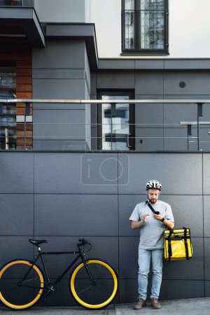 Téléchargez les photos : Jeune messager express de livraison de nourriture avec sac isolé sur le côté regarde son téléphone près du vélo garé. - en image libre de droit