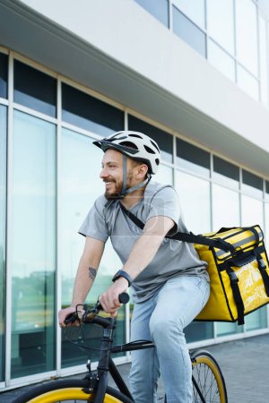Téléchargez les photos : Jeune sourire messager express livraison de nourriture est sur un vélo avec un sac isolé derrière son dos. - en image libre de droit