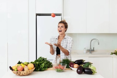 Foto de Mujer alegre está lanzando tomate rojo durante la cocina comida vegetariana en casa - Imagen libre de derechos