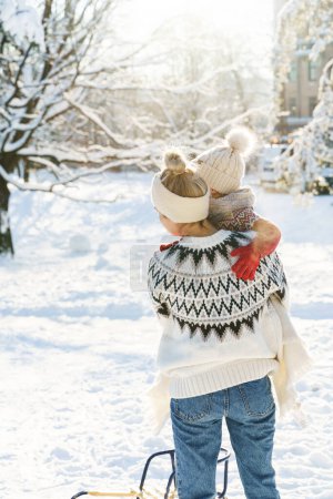 Téléchargez les photos : Jeune mère et son mignon petit fils avec traîneau vintage dans un parc enneigé pendant la journée ensoleillée d'hiver - en image libre de droit
