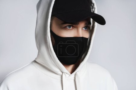 Photo for Young man wearing white hoodie, black baseball cap and cloth face mask against gray background - Royalty Free Image
