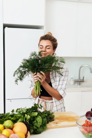 Téléchargez les photos : Jeune jolie femme avec un gros bouquet d'aneth vert pendant la cuisson dans la cuisine - en image libre de droit