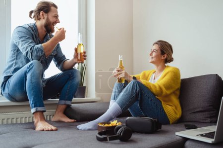 Foto de Joven pareja alegre sentada en el sofá bebiendo cerveza y comiendo nachos en casa - Imagen libre de derechos