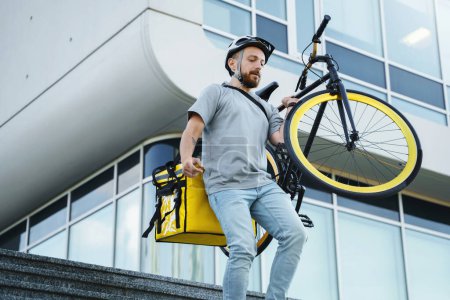 Foto de Joven mensajero de reparto de comida express con bolsa aislada detrás de su espalda está llevando su bicicleta por las escaleras. - Imagen libre de derechos