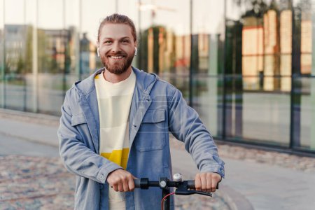 Téléchargez les photos : Portrait de jeune homme joyeux chevauchant son e-scooter dans la rue de la ville - en image libre de droit